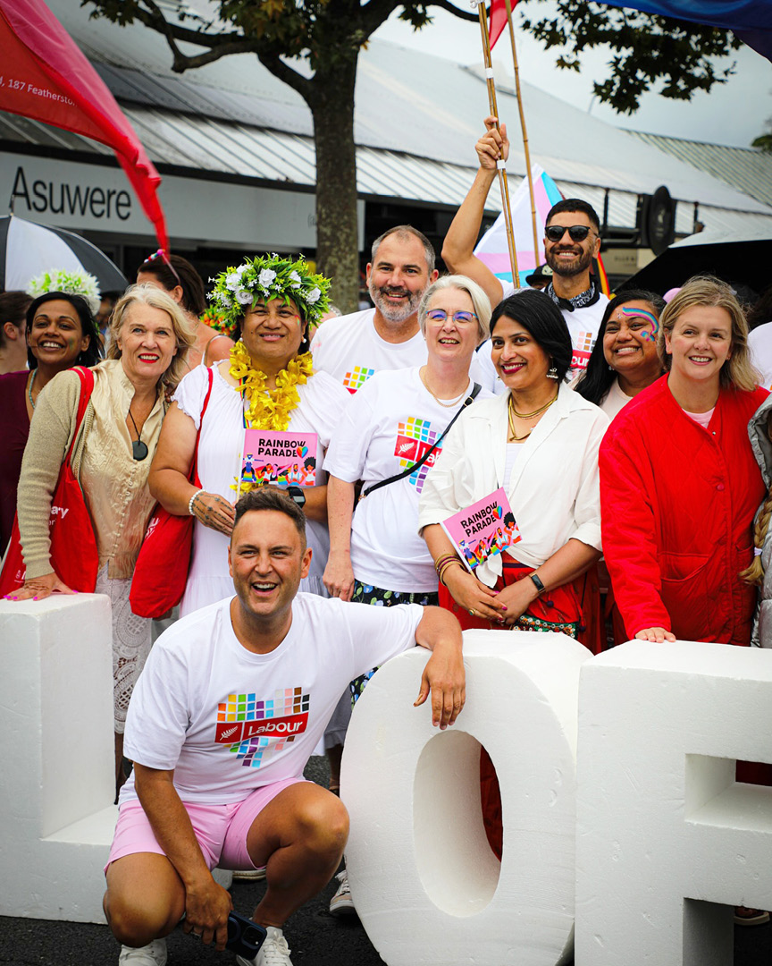Labour MPs including Chris Hipkins, Shanan Halbert, Carmel Sepuloni, and Willow-Jean Prime standing in front of the 'All of us' signage at the Auckland Pride event in 2025, wearing the Labour Pride t-shirts designed by Bradley Pratt.